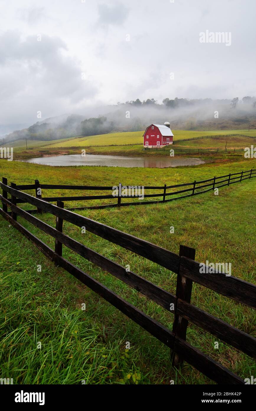 A bright red barn sits in stark contrast against a green landscape framed by hills, a small farm pond and fence line on a foggy, raining day near Marl Stock Photo