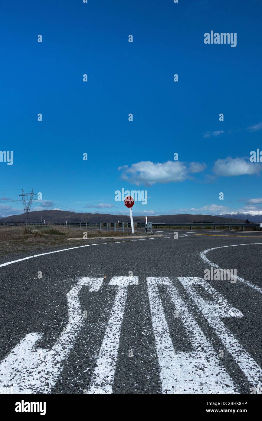 Large 'stop' lettering painted on tarmac road & red sign post viewed from low level with blue sky, clouds & snow capped mountains in background Stock Photo