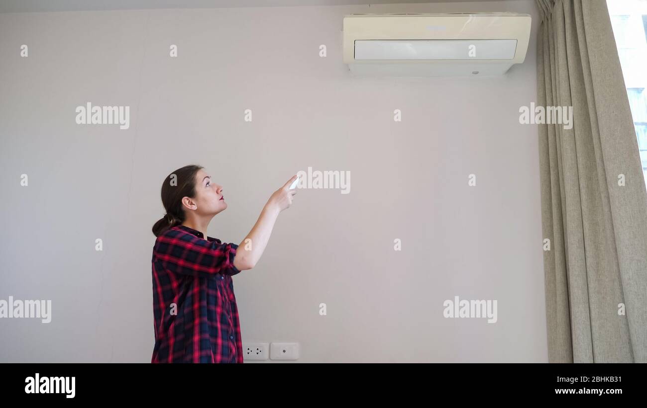brunette with long hair in ponytail turns on air conditioner with remote control and smiles standing near white wall Stock Photo