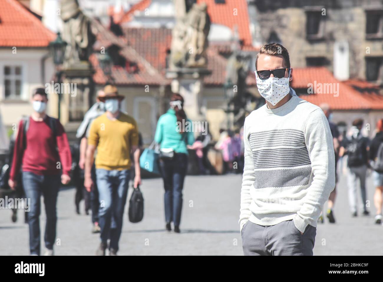 Young man with sunglasses and sewed fabric face mask photographed on the Charles Bridge in Prague, Czech Republic. Blurred people in the background. Travelling, tourism during coronavirus. COVID-19. Stock Photo