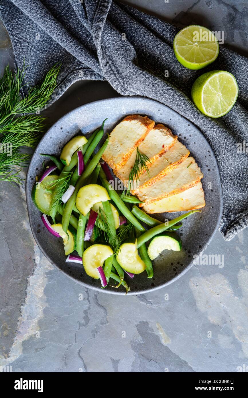 Marinated tofu slices served with blanched green beans and zucchini, drizzled with olive oil and decorated with fresh dill on a ceramic plate Stock Photo