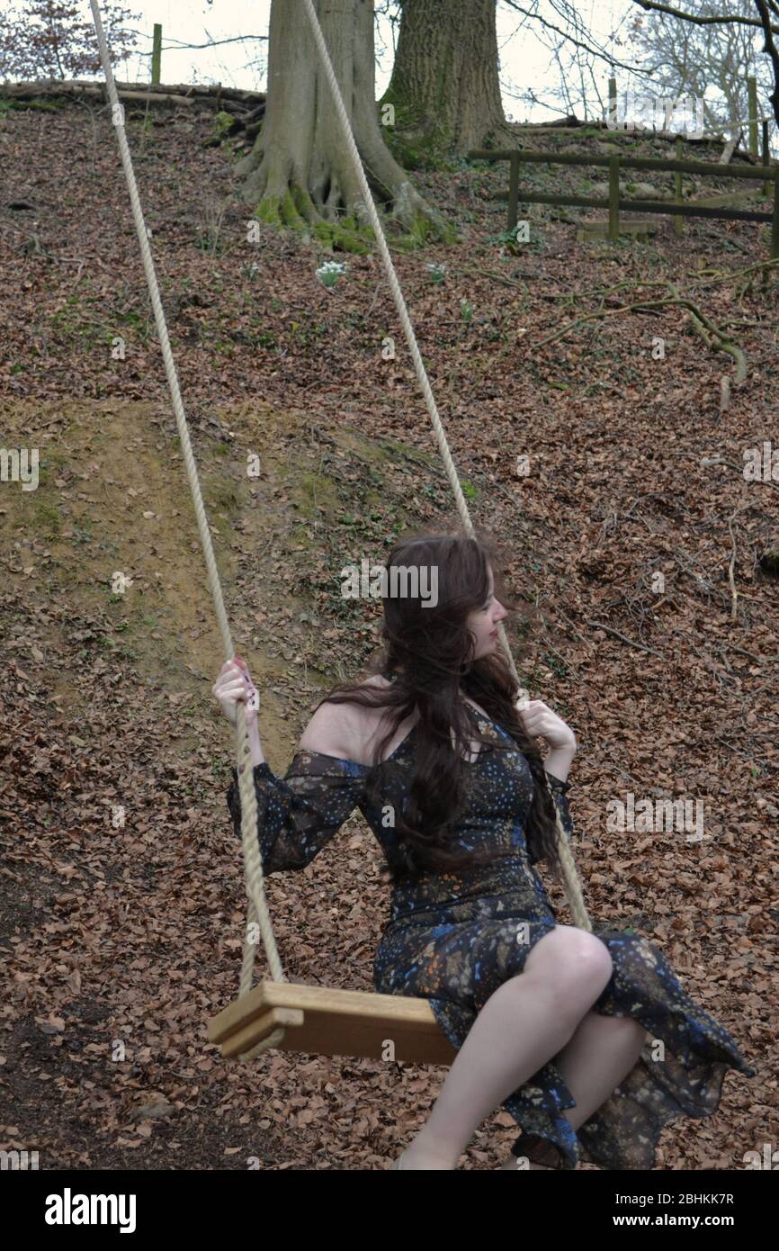 A long haired brunette young lady in a formal dress playing on a swing in winter woodland Stock Photo