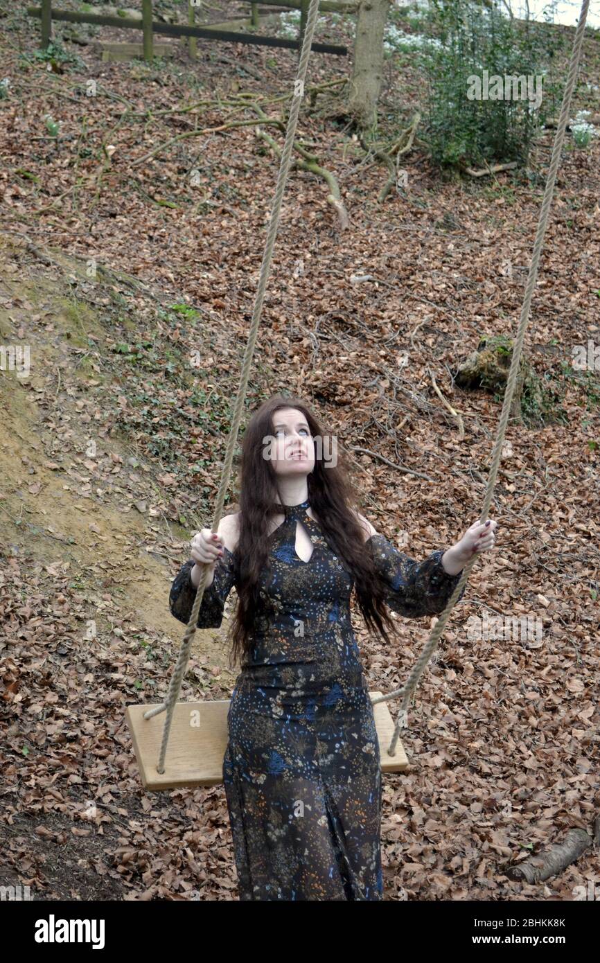 A long haired brunette young lady in a formal dress playing on a swing in winter woodland, nervously testing the ropes Stock Photo