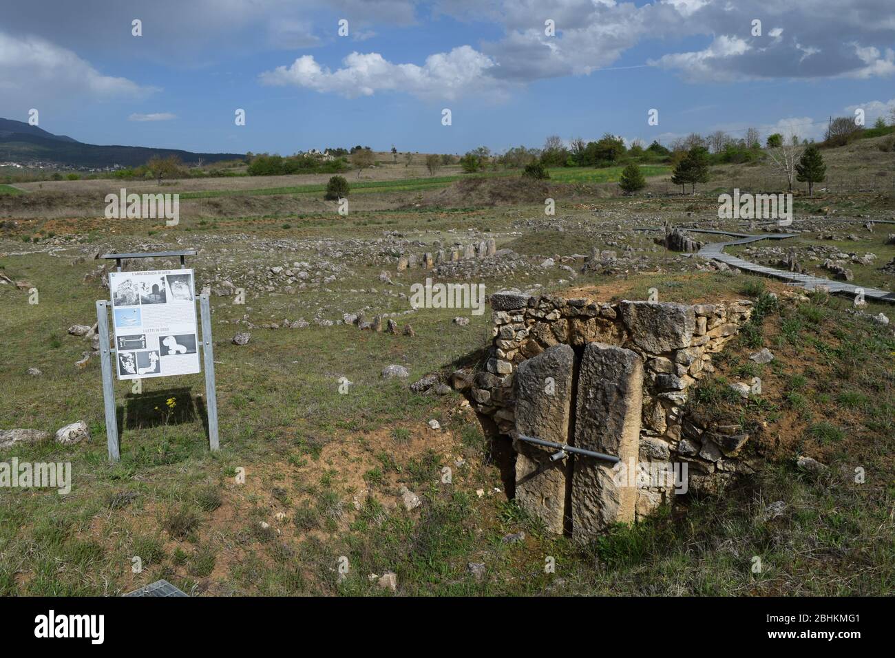  An image of a large, grassy field containing two large stone structures with stone lintels, one being a doorway, and the other having a large stone slab leaning against it, with a small sign in the foreground describing the ruins in Italian.