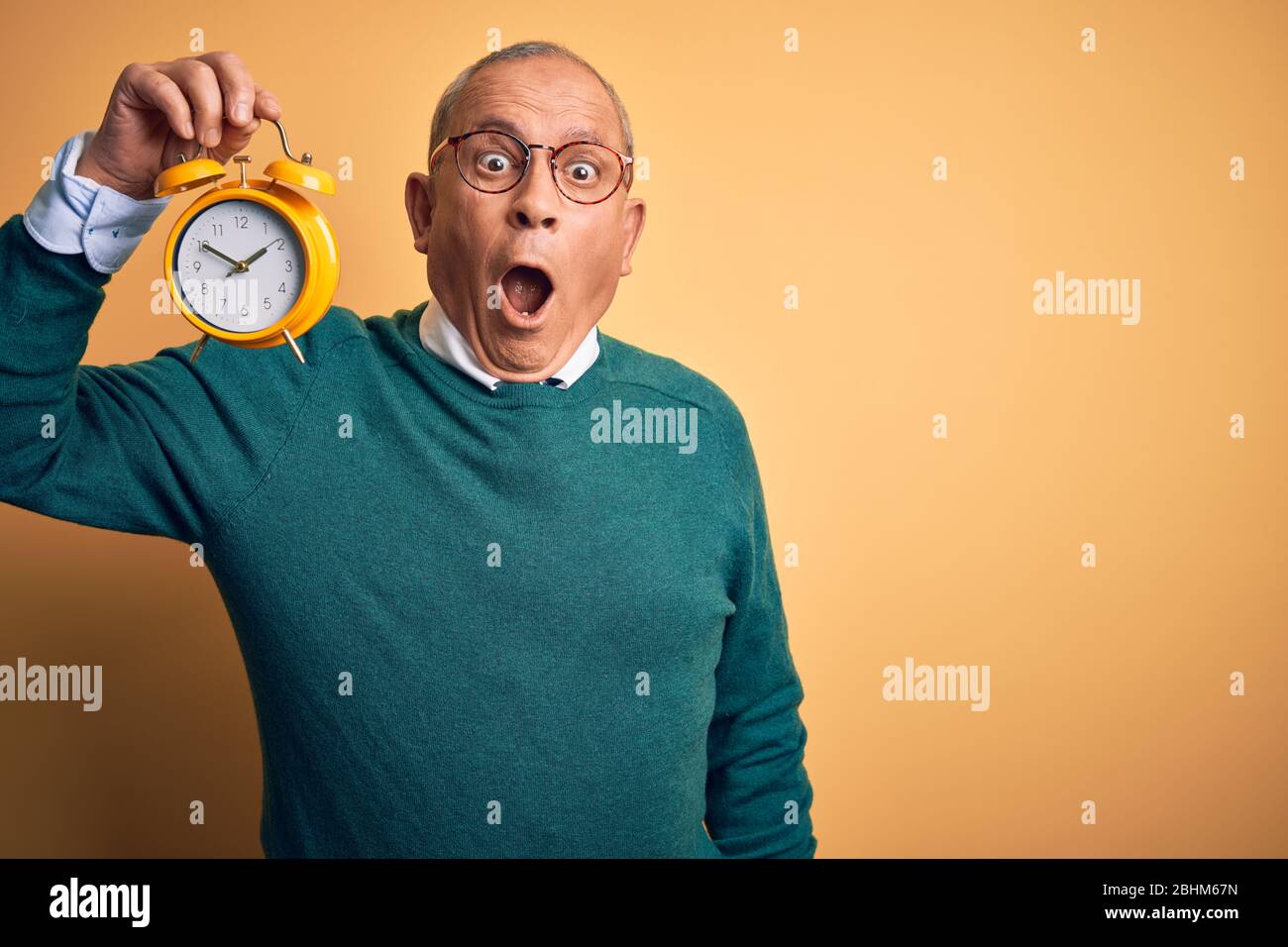 Senior handsome man holding alarm clock standing over isolated yellow