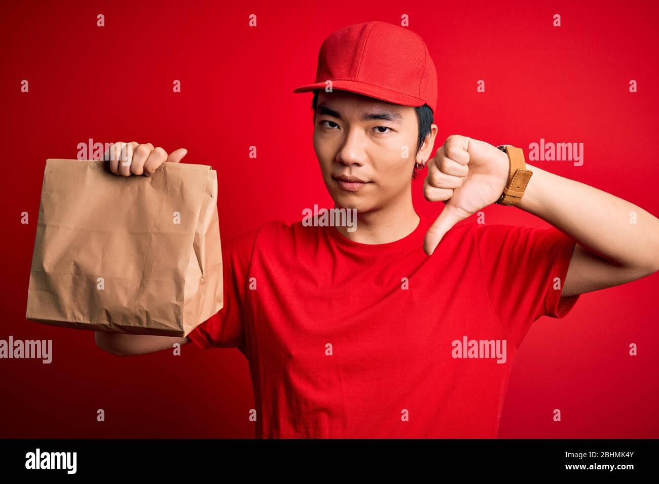 Young handsome chinese delivery man holding takeaway paper bag with food with angry face