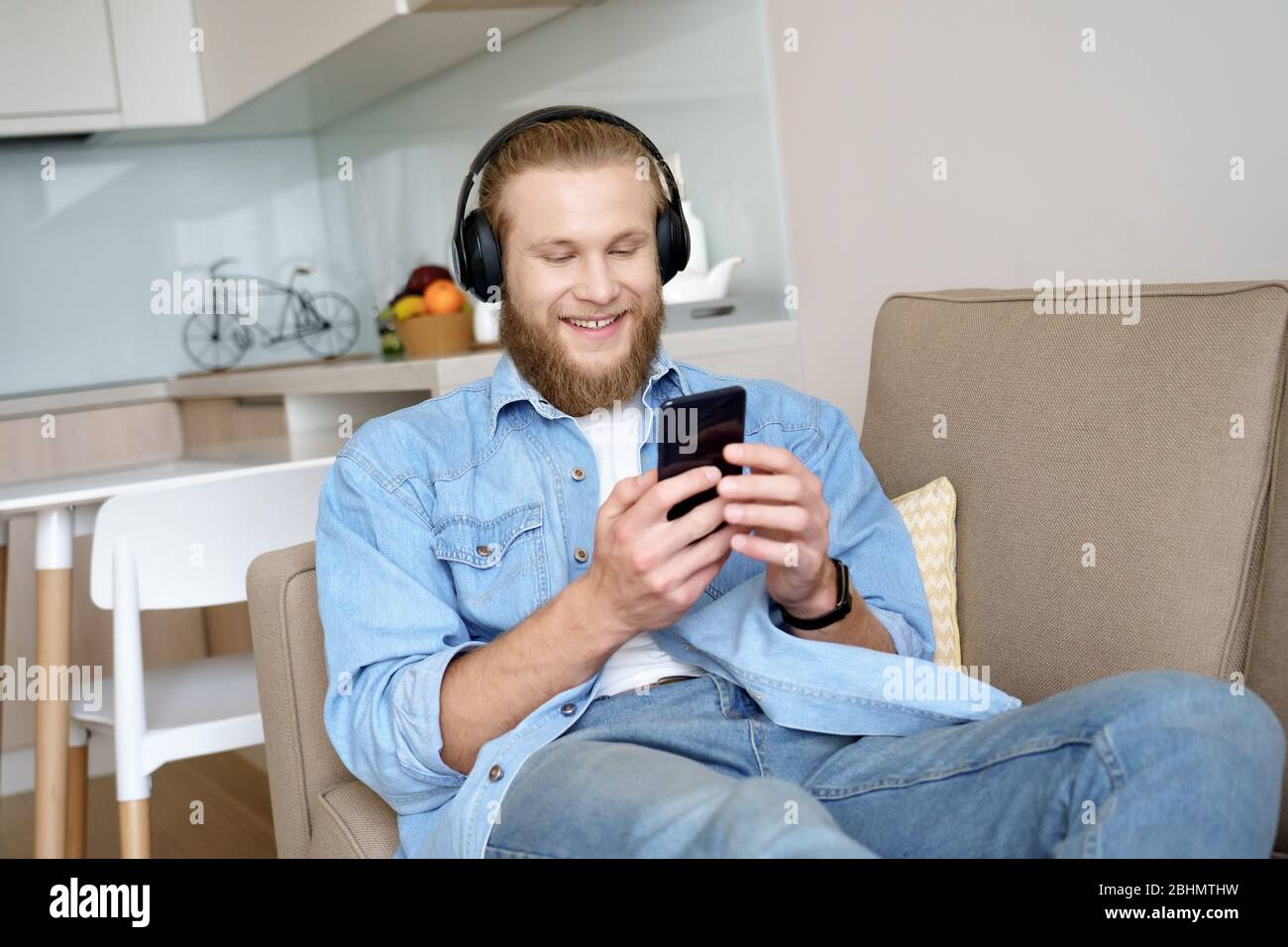 Happy young guy wearing headphones using phone relax on sofa. Stock Photo