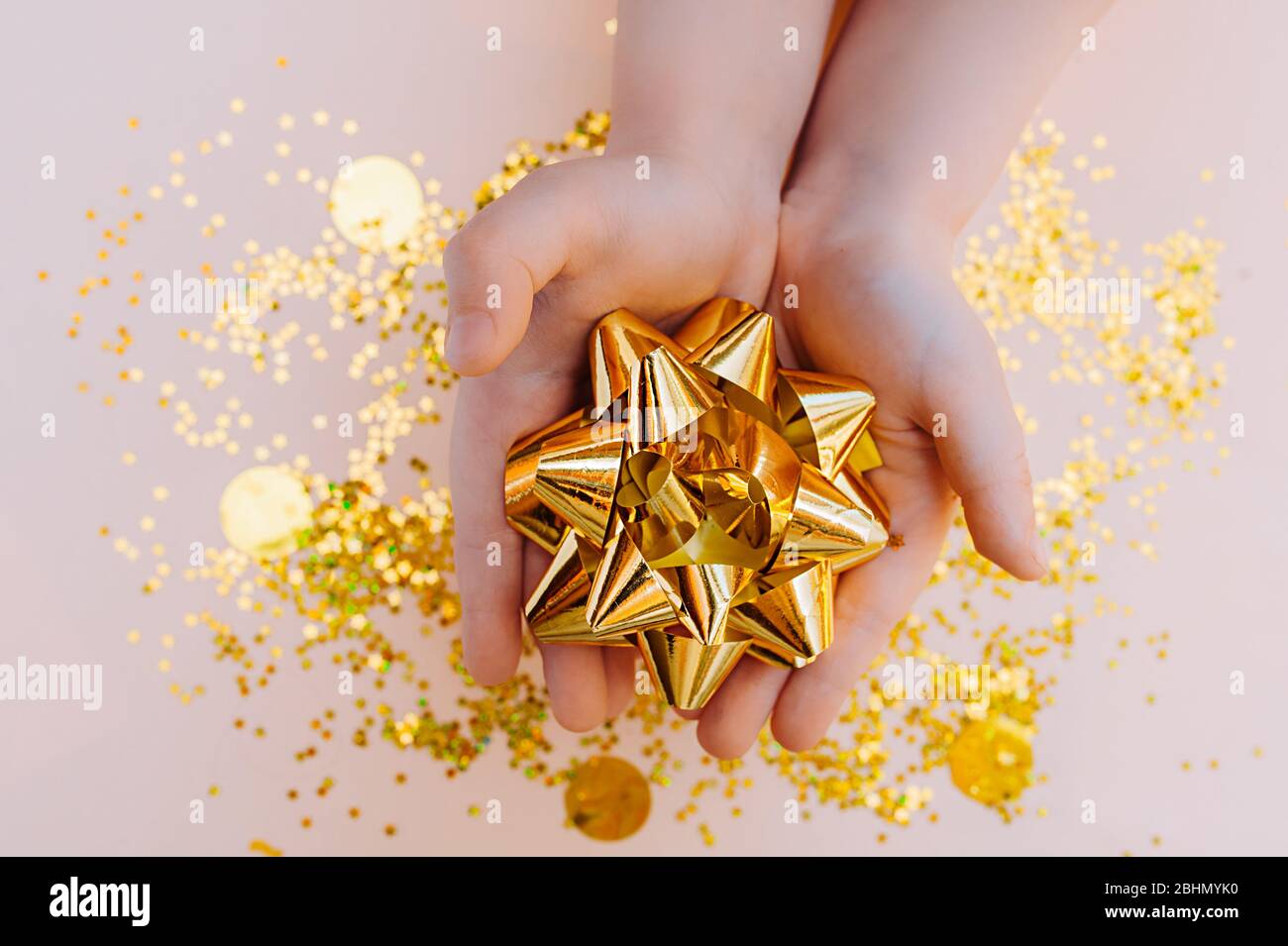 Children's hands light skin holding a large gift bow made of gold foil on a background of scattered gold sequins Stock Photo