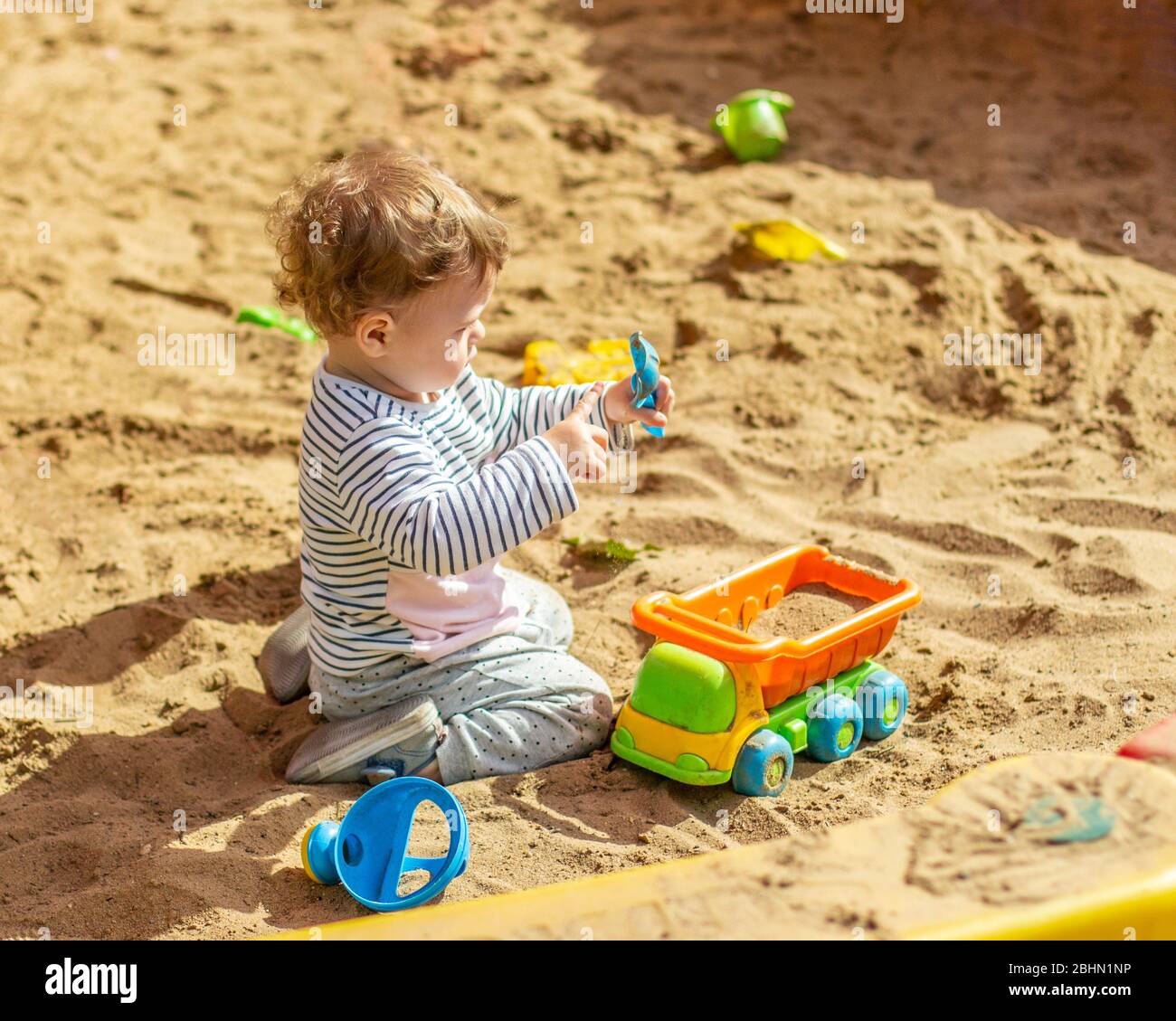 Little baby girl Caucasian boy plays in the sandbox with a toy car. A child in a striped t-shirt pours sand into an orange truck with a plastic spatul Stock Photo