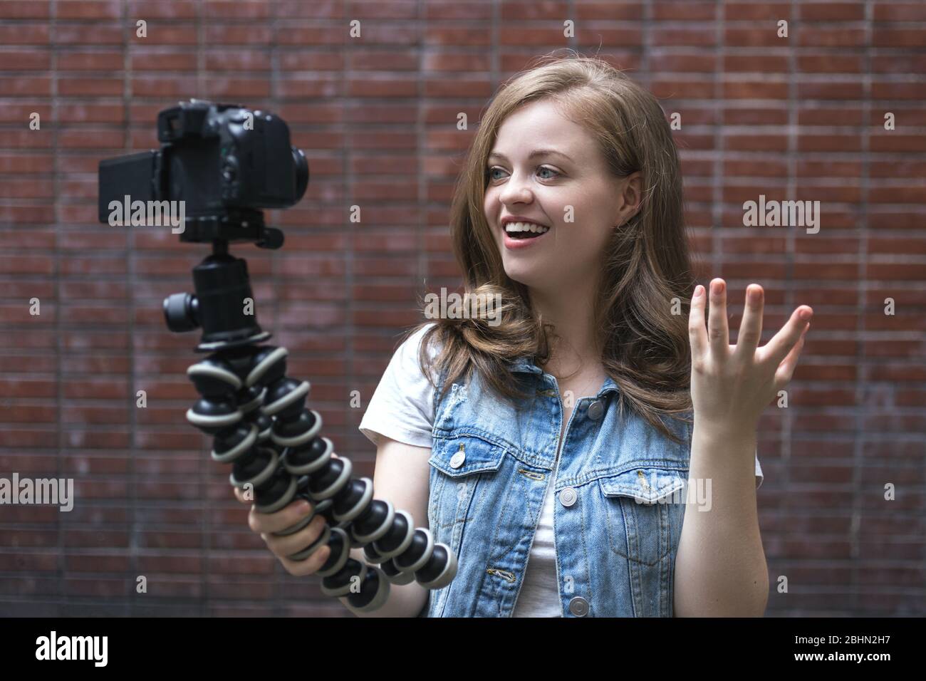 Smiling young caucasian girl woman making a video blog (vlog) with camera near brick wall Stock Photo