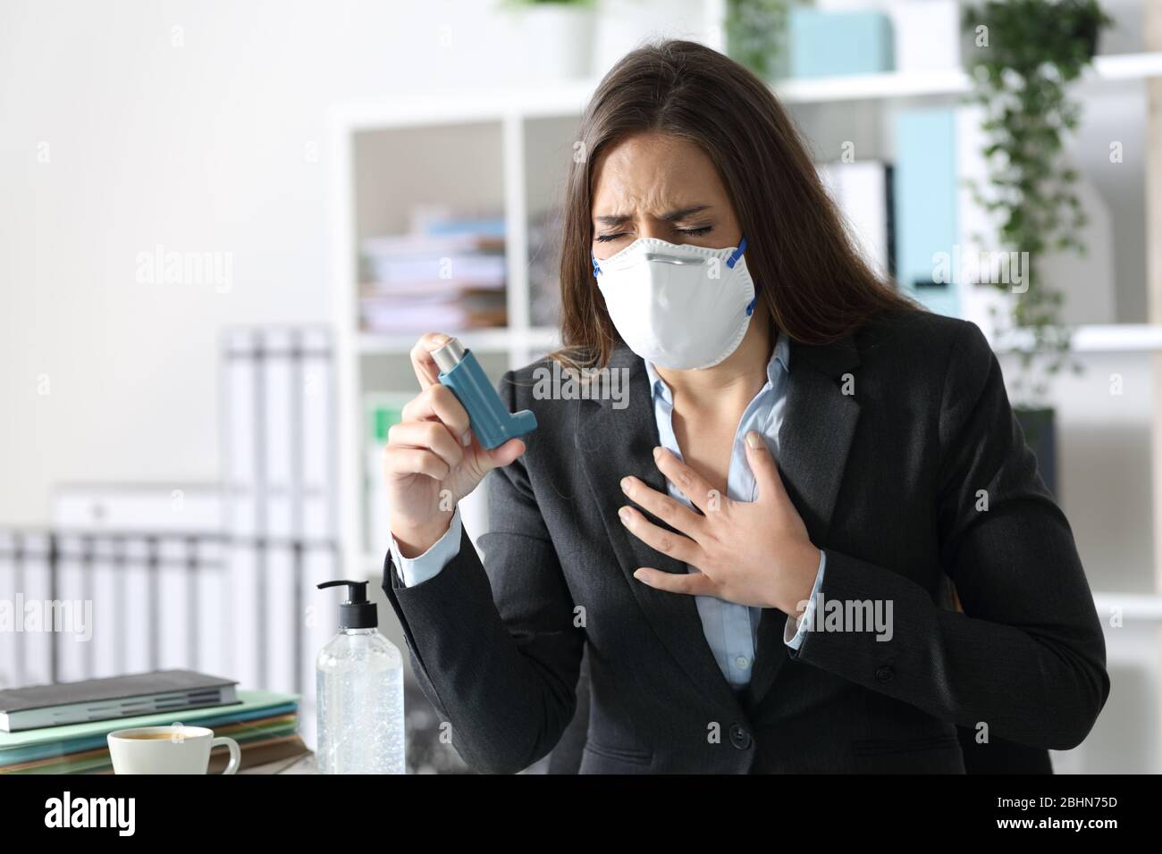 Executive woman wearing protective mask with asthma attack holding inhaler touching chest at the office Stock Photo