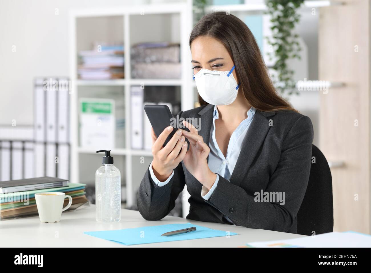 Executive woman with protective mask using smart phone sitting on a desk at the office Stock Photo