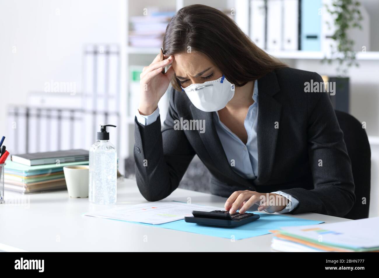 Worried bookkeeper with protective mask looking at calculator on a desk at the office Stock Photo