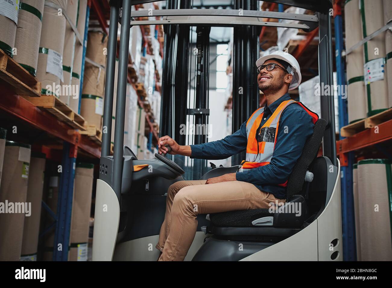 African forklift driver focused on carefully transporting stock from shelves of a large warehouse wearing a white helmet and vest looking up towards Stock Photo