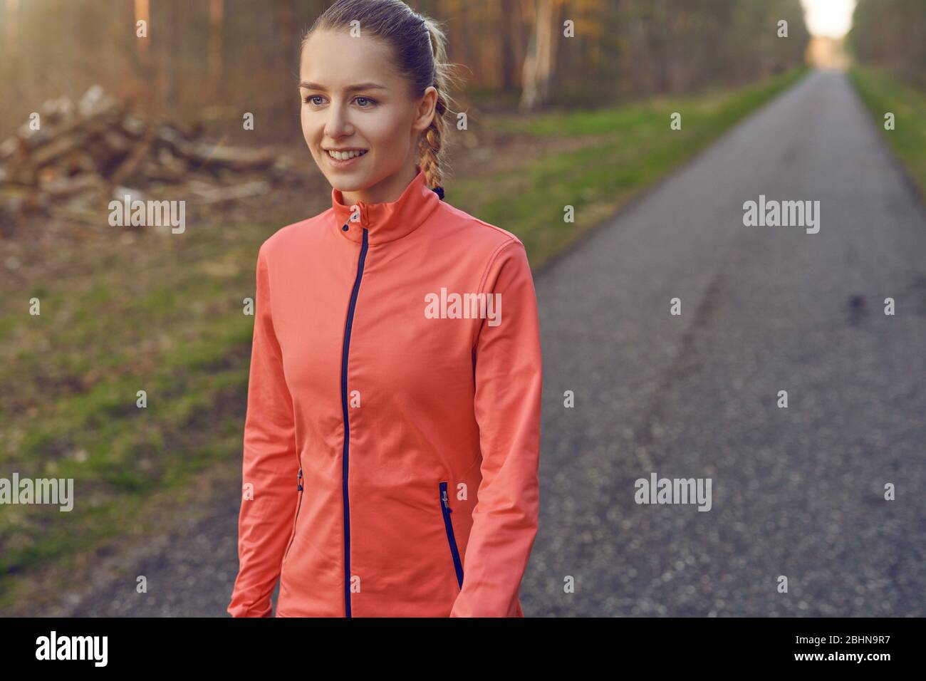 Smiling athletic fit young woman working out on a tarred lane through forests backlit by the warm glow of the sun in a healthy active lifestyle concep Stock Photo