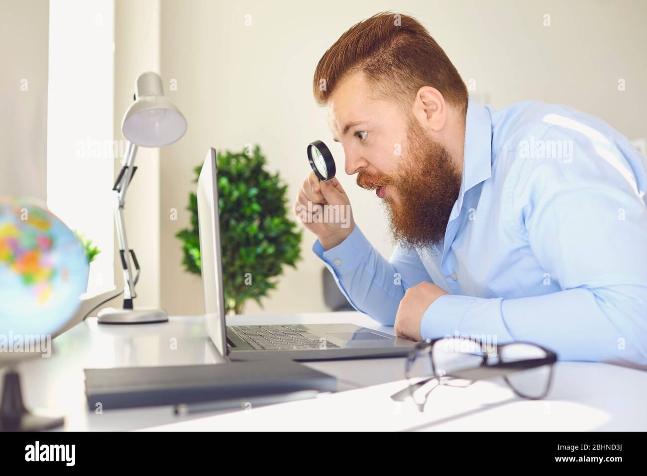 Online work at home. A funny fat bearded man with a laptop increases magnifying information at a table at home. Stock Photo