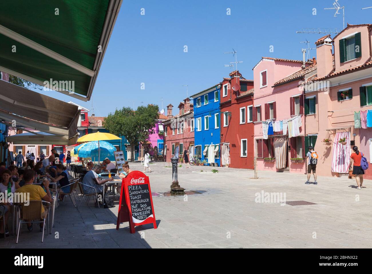 Burano, Venice, Veneto, Italy. Tourists eating at an outdoor pizzeria at lunchtime in summer  overlooking colourful houses in Corte della Comare Stock Photo