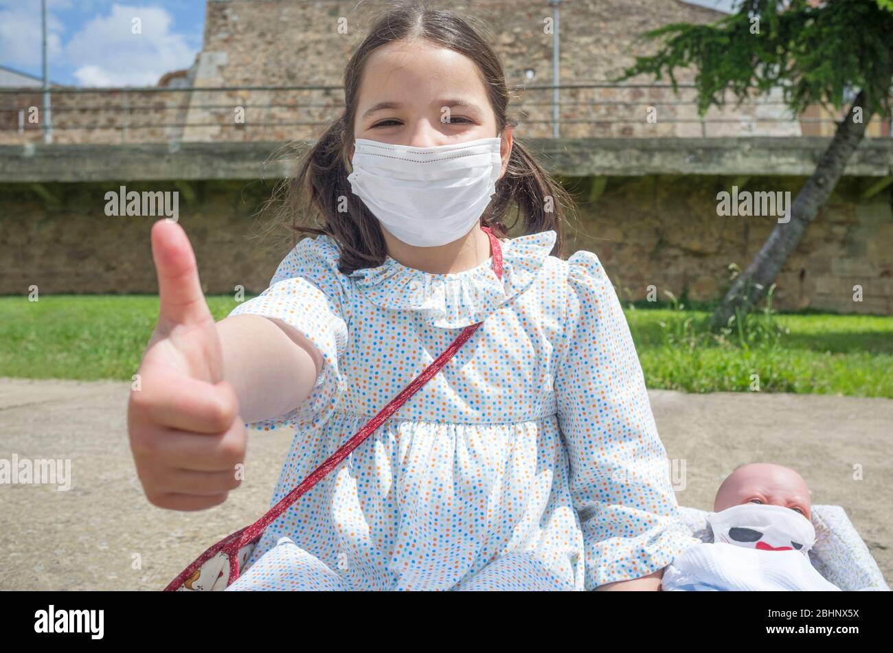 Little girl happy in her first day of children outing cause relaxation of confinement measures. SHe and her reborn doll are wearing face mask Stock Photo