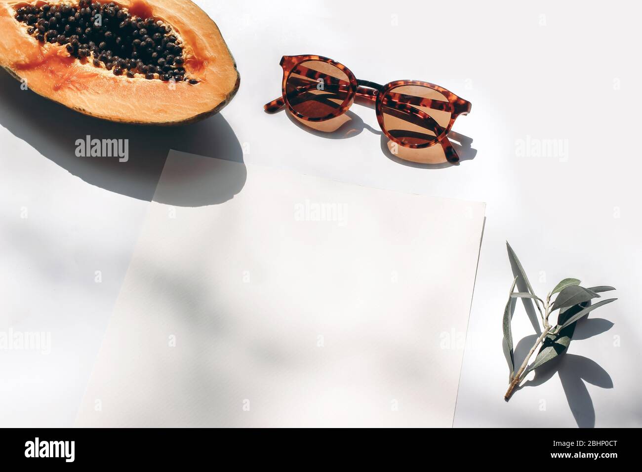 Summer stationery still life scene. Closeup of cut papaya fruit, olive branch and sunglasses in sunlight. White table. Blank paper cards, invitations Stock Photo