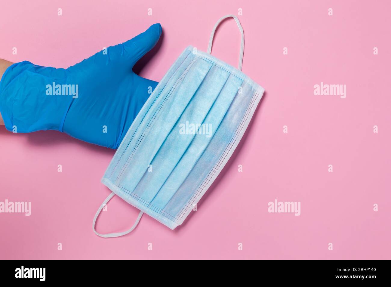 Coronavirus protection. Hand in blue disposable latex glove holding medical surgical mask on pink background. Hygiene measures to prevent spread of Co Stock Photo