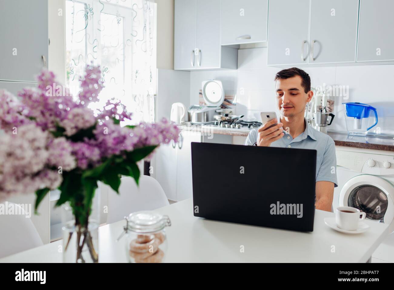 Young businessman working from home during coronavirus lockdown using laptop and smartphone having coffee. Stock Photo