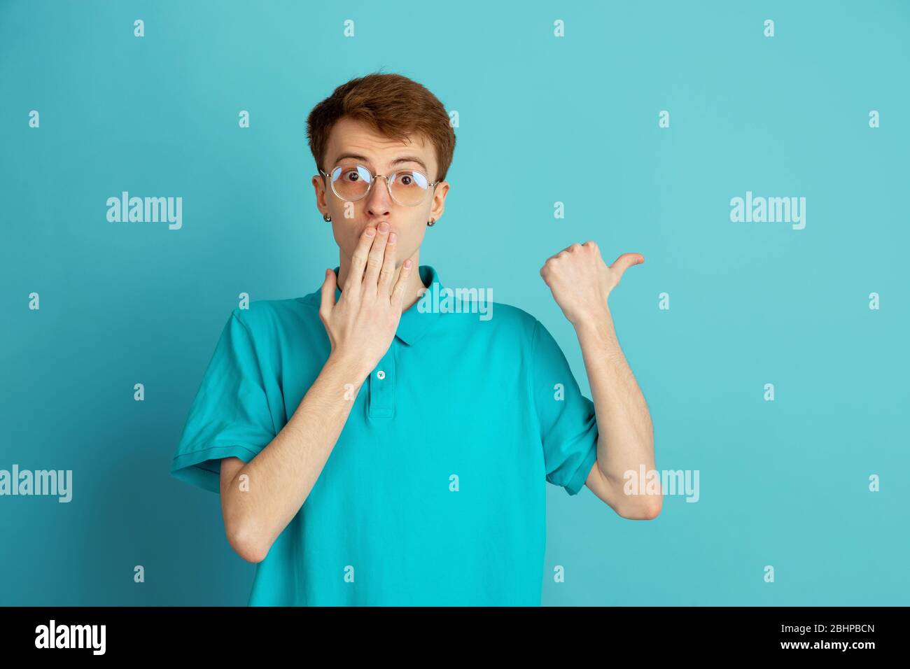 Pointing at side. Caucasian young man's modern portrait isolated on blue studio background, monochrome. Beautiful male model. Concept of human emotions, facial expression, sales, ad, trendy. Stock Photo