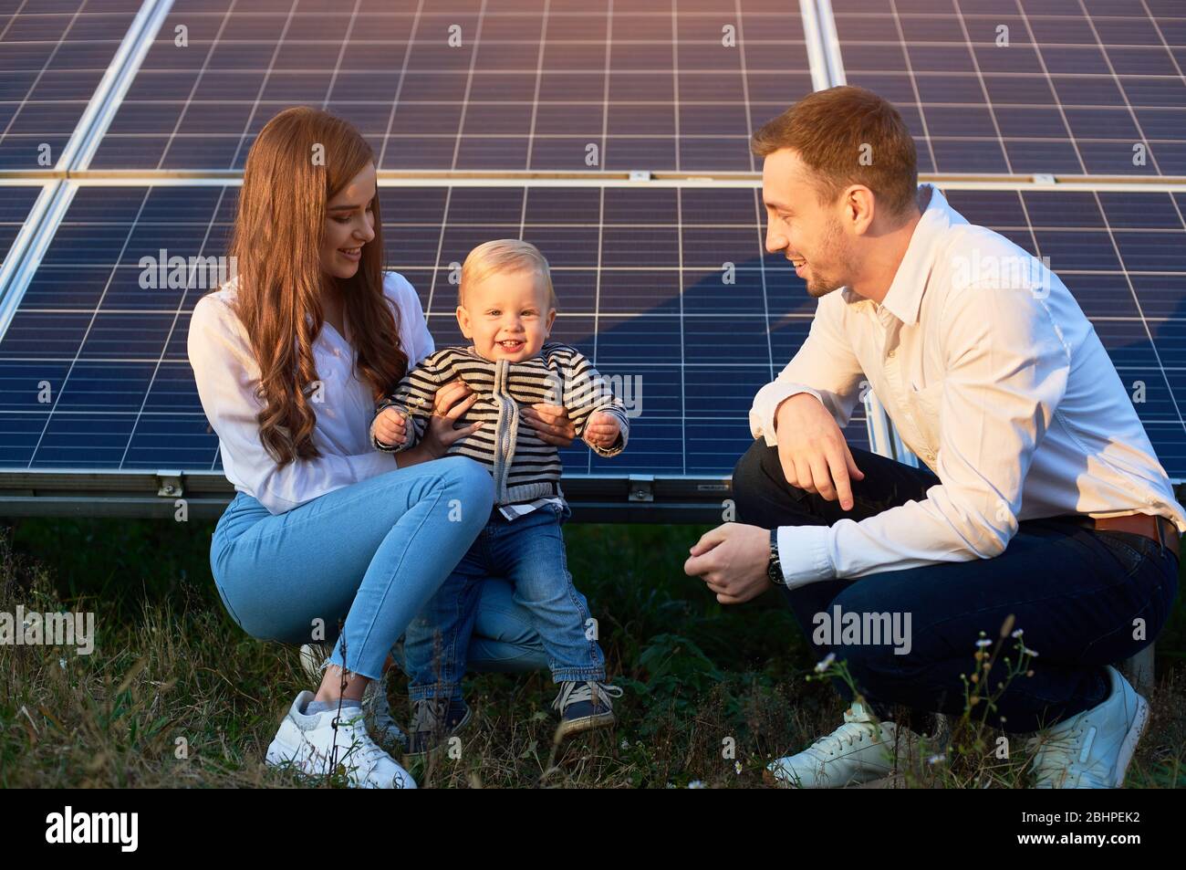 Young family of three is crouching near photovoltaic solar panel, little boy is looking at camera, parents looking at him, modern family concept Stock Photo