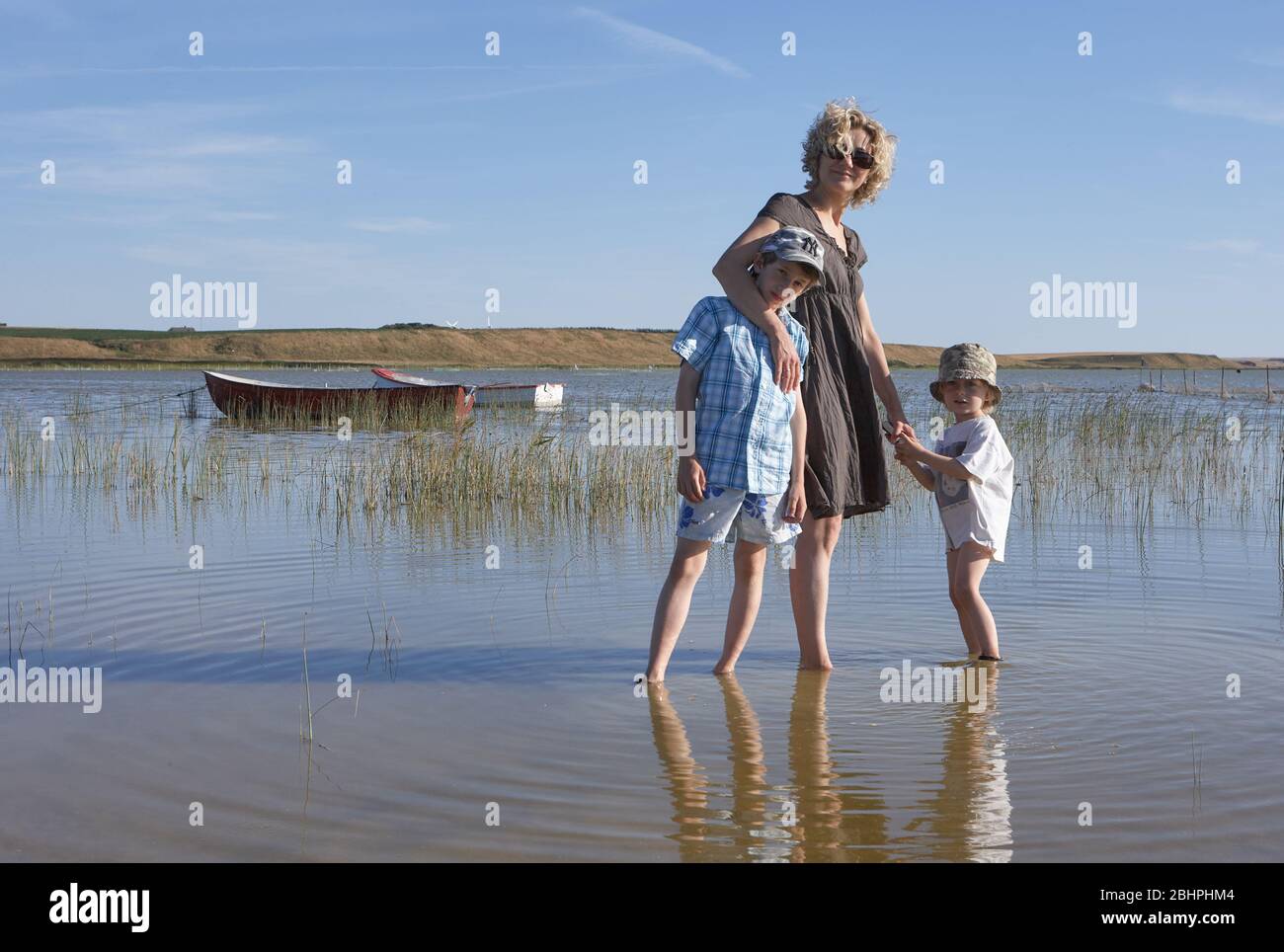 Blond mother with sunglasses, holding the sands of her son while keeping the other one in her arm, walking around in a lake with reed and boats Stock Photo