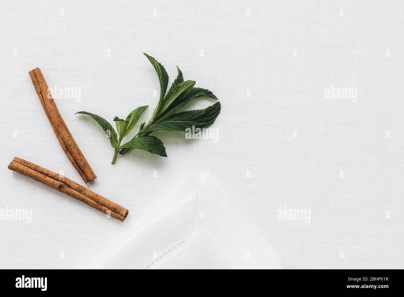 Fresh mint and fragrant cinnamon sticks on a white linen tablecloth in the morning light. Stock Photo