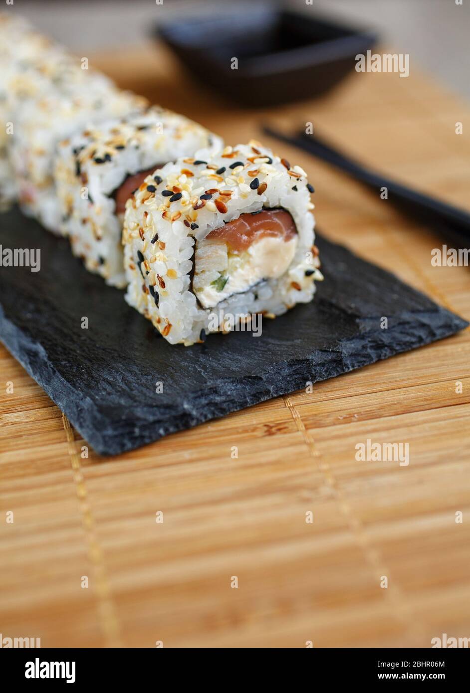 Uramaki Maki Sushi, Original roll served on a black board with black chopsticks and soy sauce in a black bowl. Stock Photo