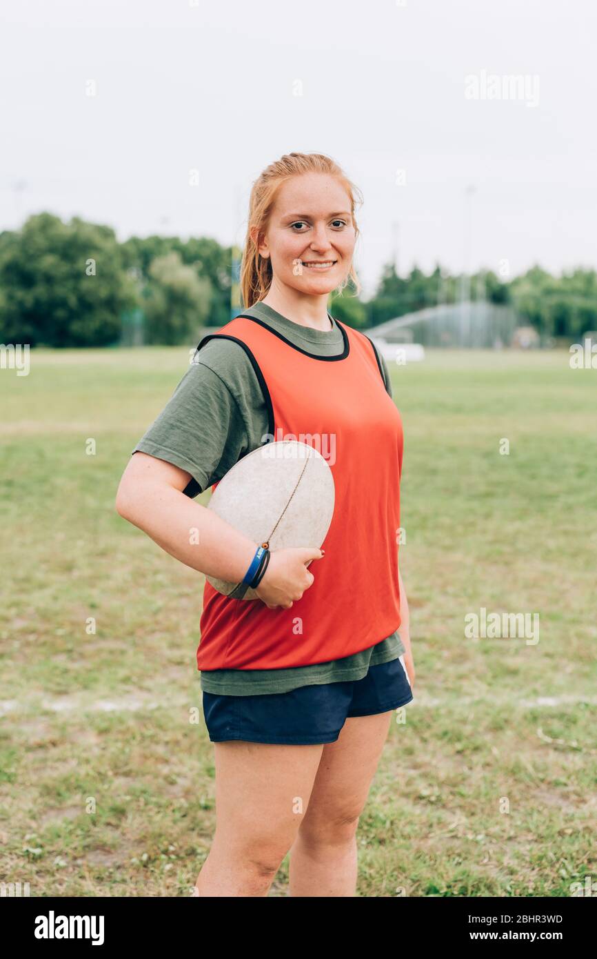A woman standing on a training pitch in shorts and tee shirt holding a rugby ball. Stock Photo