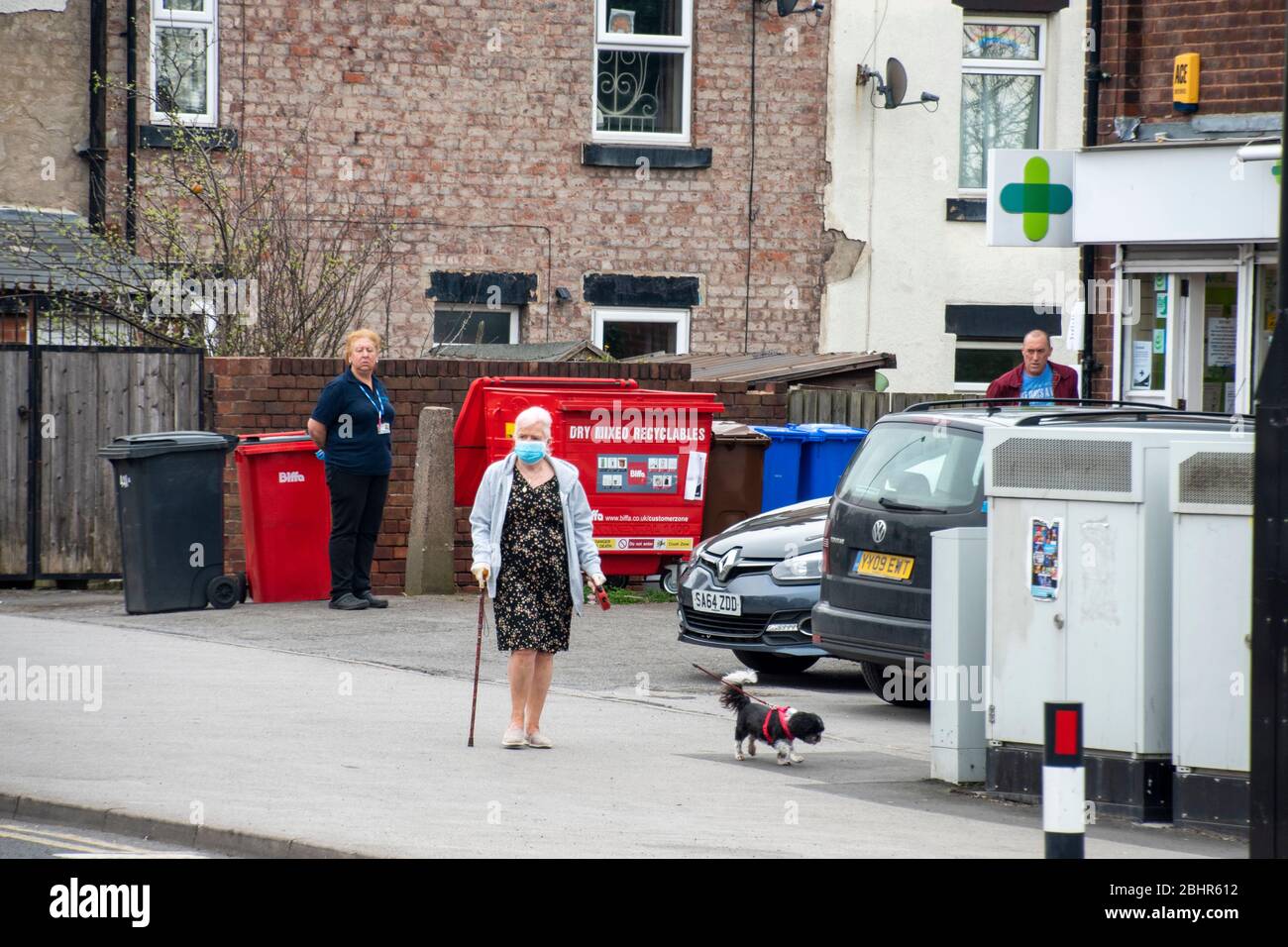 Sheffield UK –  March 30 2020: Elderly woman in PPE mask walks past socially distanced queue at Well Pharmacy during covid-19 pandemic, Richmond Road Stock Photo