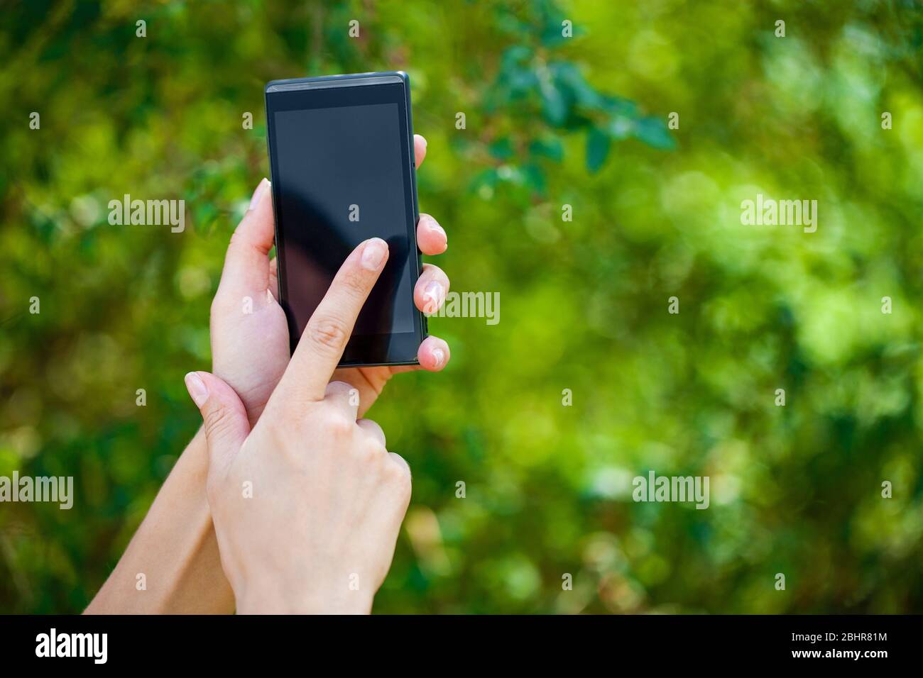Side view of a woman's hand holding a modern slick smartphone while dialing with her thumb against summer green background Stock Photo