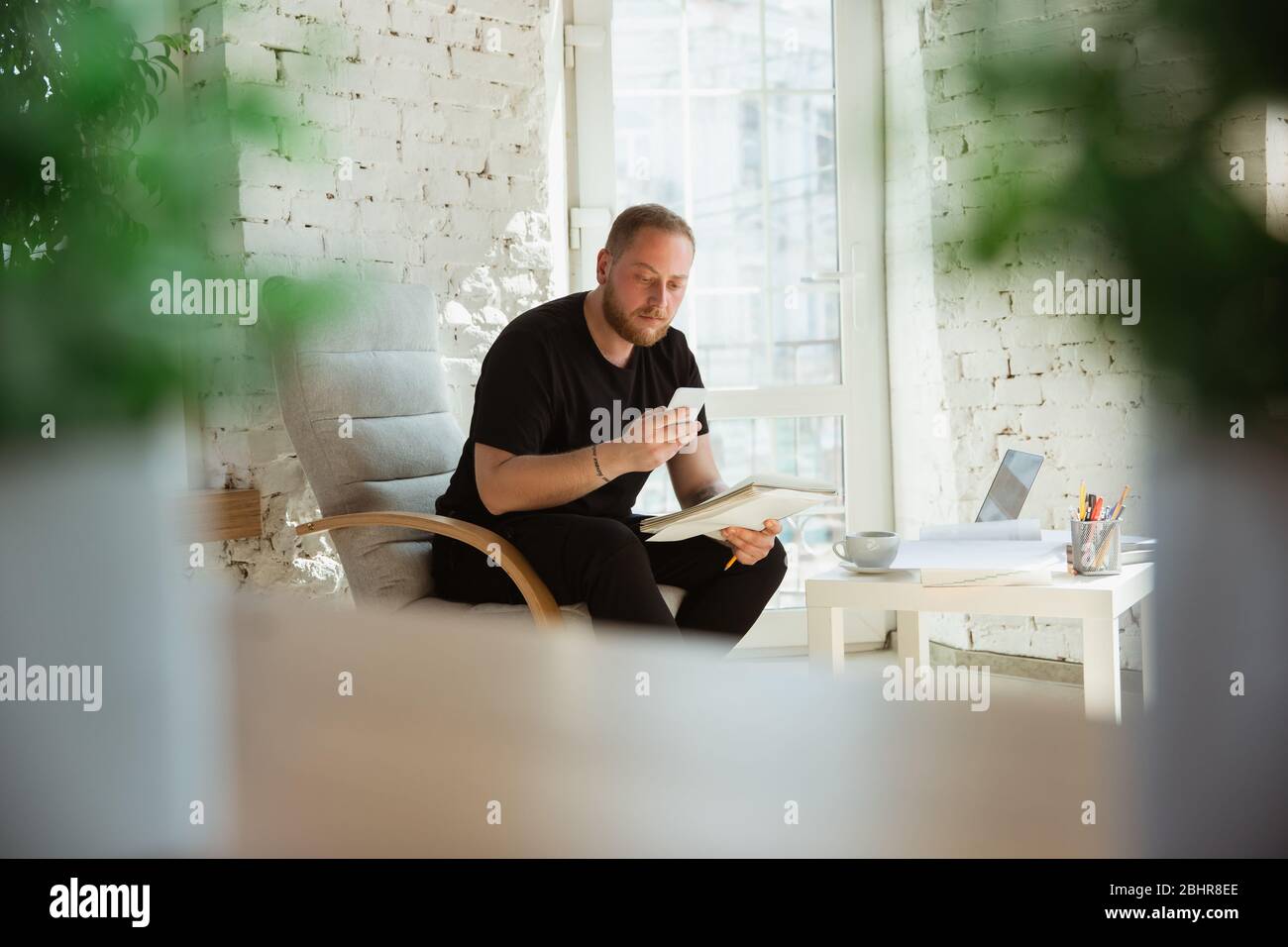 Young man studying at home during online courses for managers, marketers, buyers. Getting profession while isolated, quarantine against coronavirus spreading. Using laptop, smartphone, devices. Stock Photo