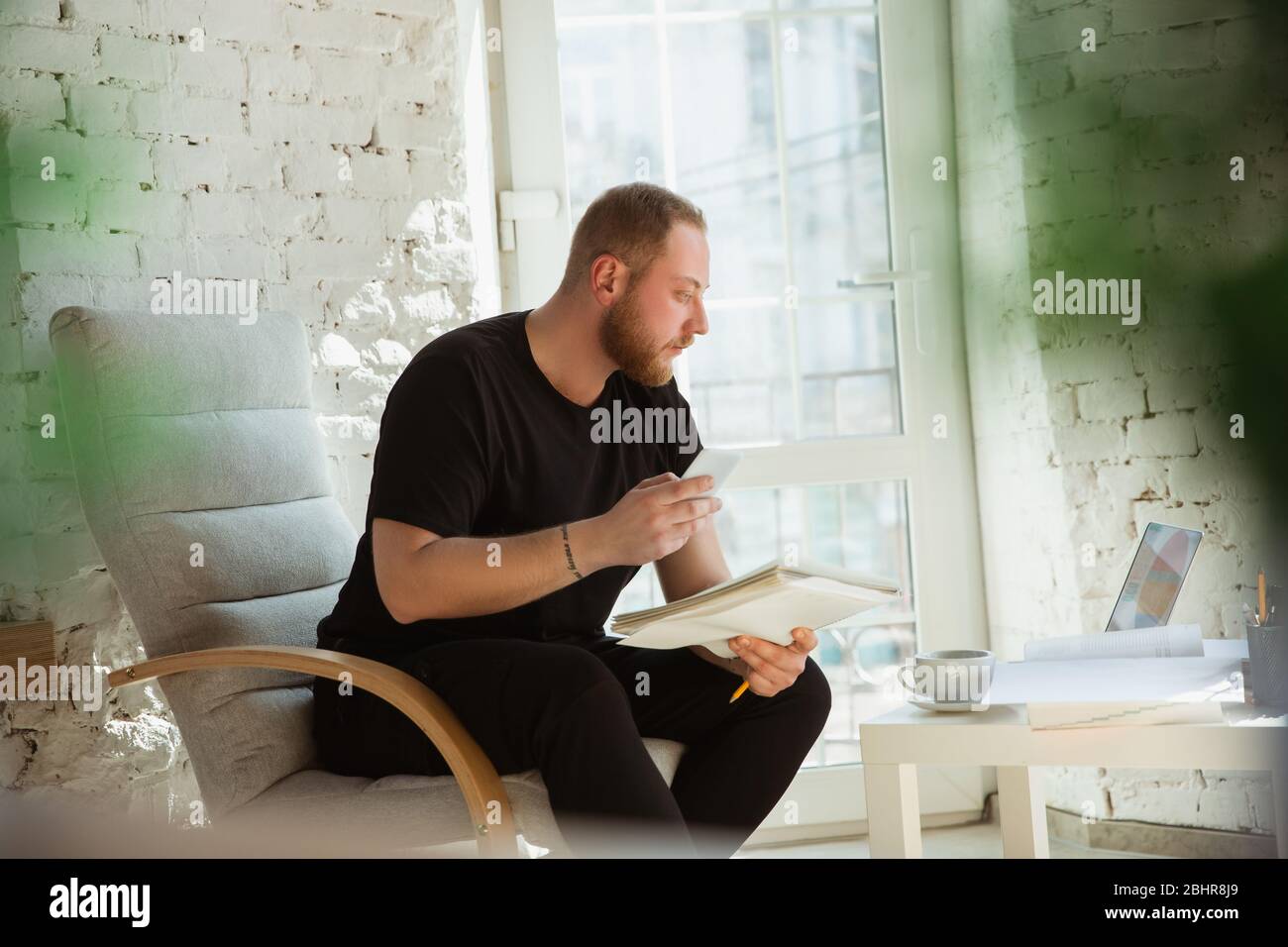 Young man studying at home during online courses for managers, marketers, buyers. Getting profession while isolated, quarantine against coronavirus spreading. Using laptop, smartphone, devices. Stock Photo