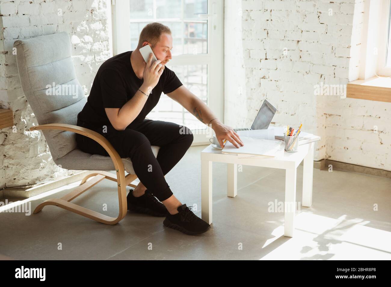 Young man studying at home during online courses for analytics, financists, economists. Getting profession while isolated, quarantine against coronavirus spreading. Using laptop, smartphone, devices. Stock Photo