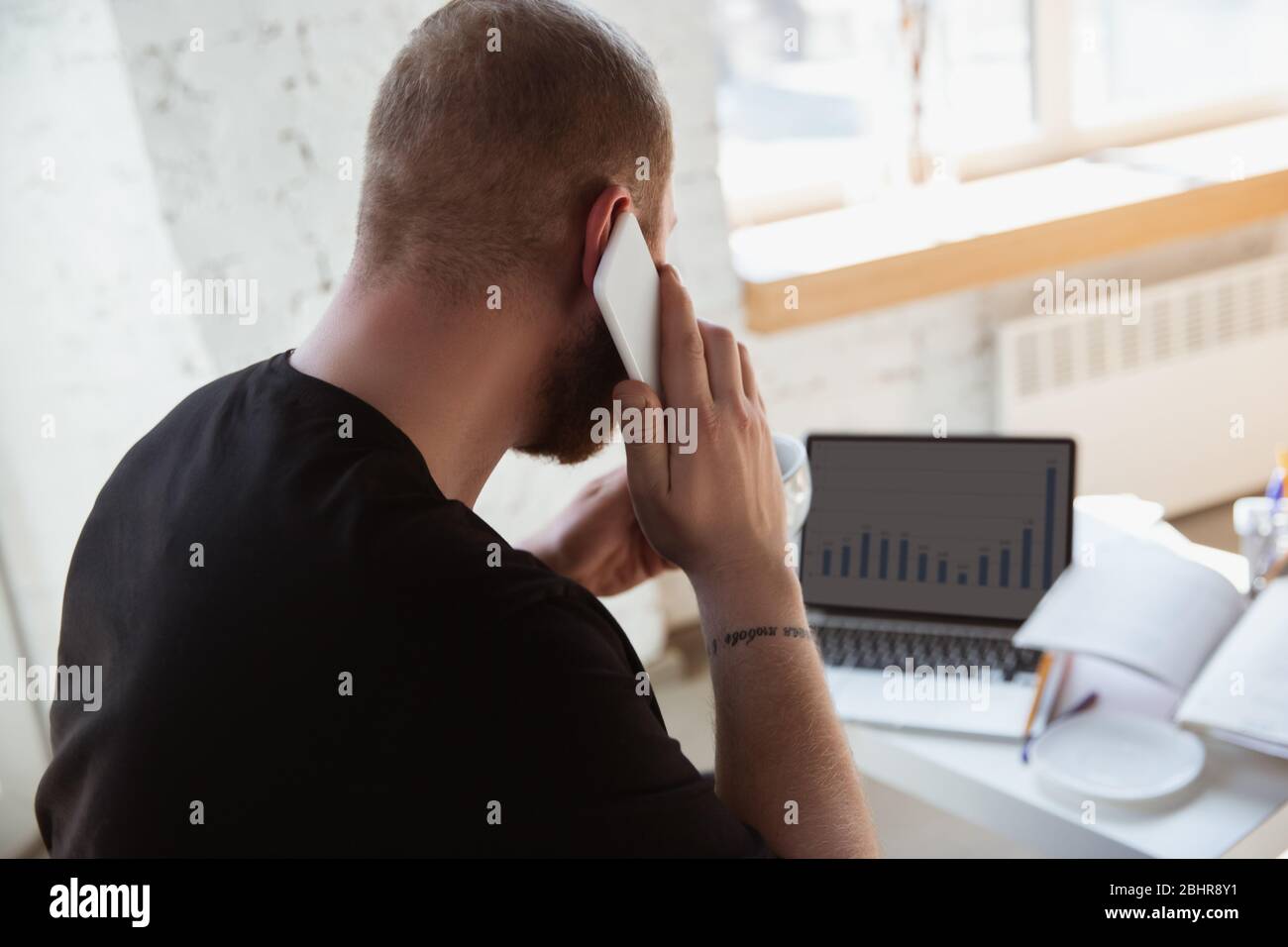 Young man studying at home during online courses for analytics, financists, economists. Getting profession while isolated, quarantine against coronavirus spreading. Using laptop, smartphone, devices. Stock Photo