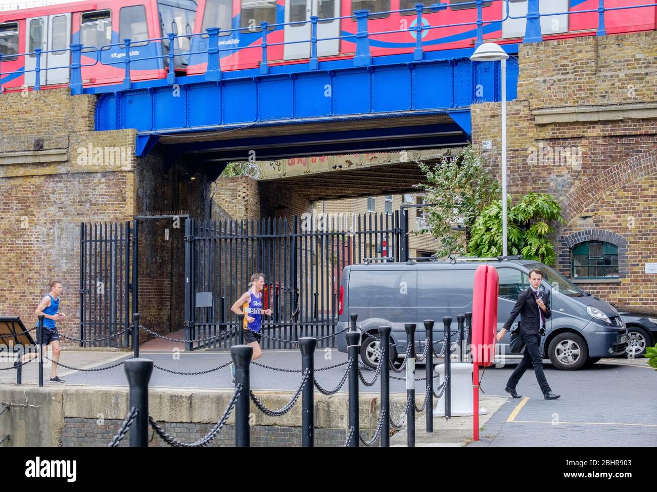 Two men jog, one man walking with suit & tie & headphones. DLR passing on bridge in background at Limehouse Marina, Tower  Hamlets, East London, UK. Stock Photo