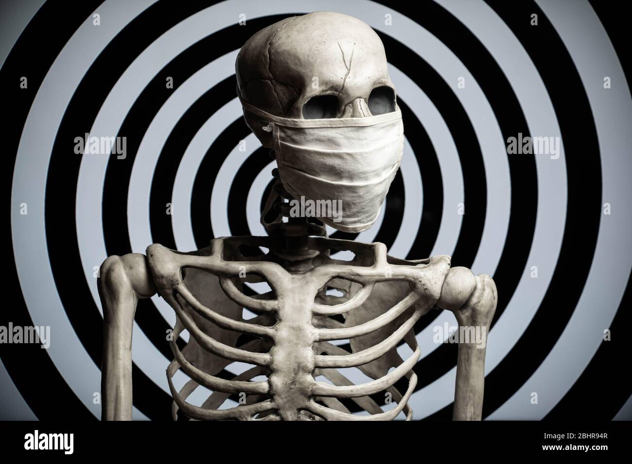 A skeleton wearing a cloth medical face mask in front of a hypnotic spiral background. Conspiracy or confusion over the covid-19 coronavirus pandemic. Stock Photo