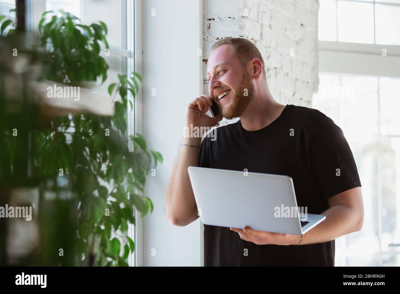 Young man studying at home during online courses for designers, consultants, IT-managers. Getting profession while isolated, quarantine against coronavirus spreading. Using laptop, smartphone, devices. Stock Photo