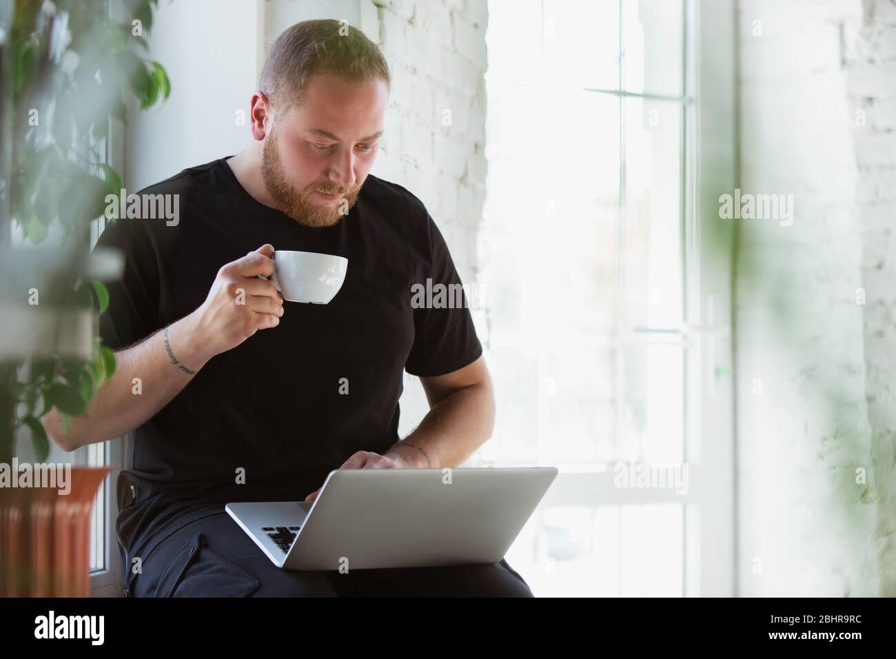 Young man studying at home during online courses for designers, consultants, IT-managers. Getting profession while isolated, quarantine against coronavirus spreading. Using laptop, smartphone, devices. Stock Photo