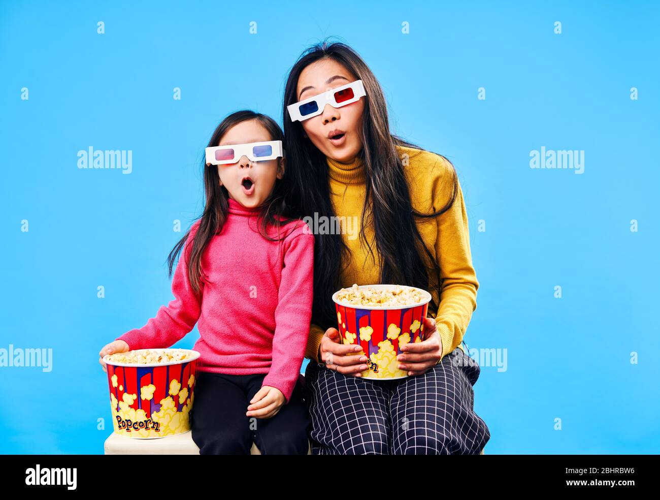 Surprised mother and her little daughter in 3D glasses with popcorn watching movie over blue background. Wow concept Stock Photo