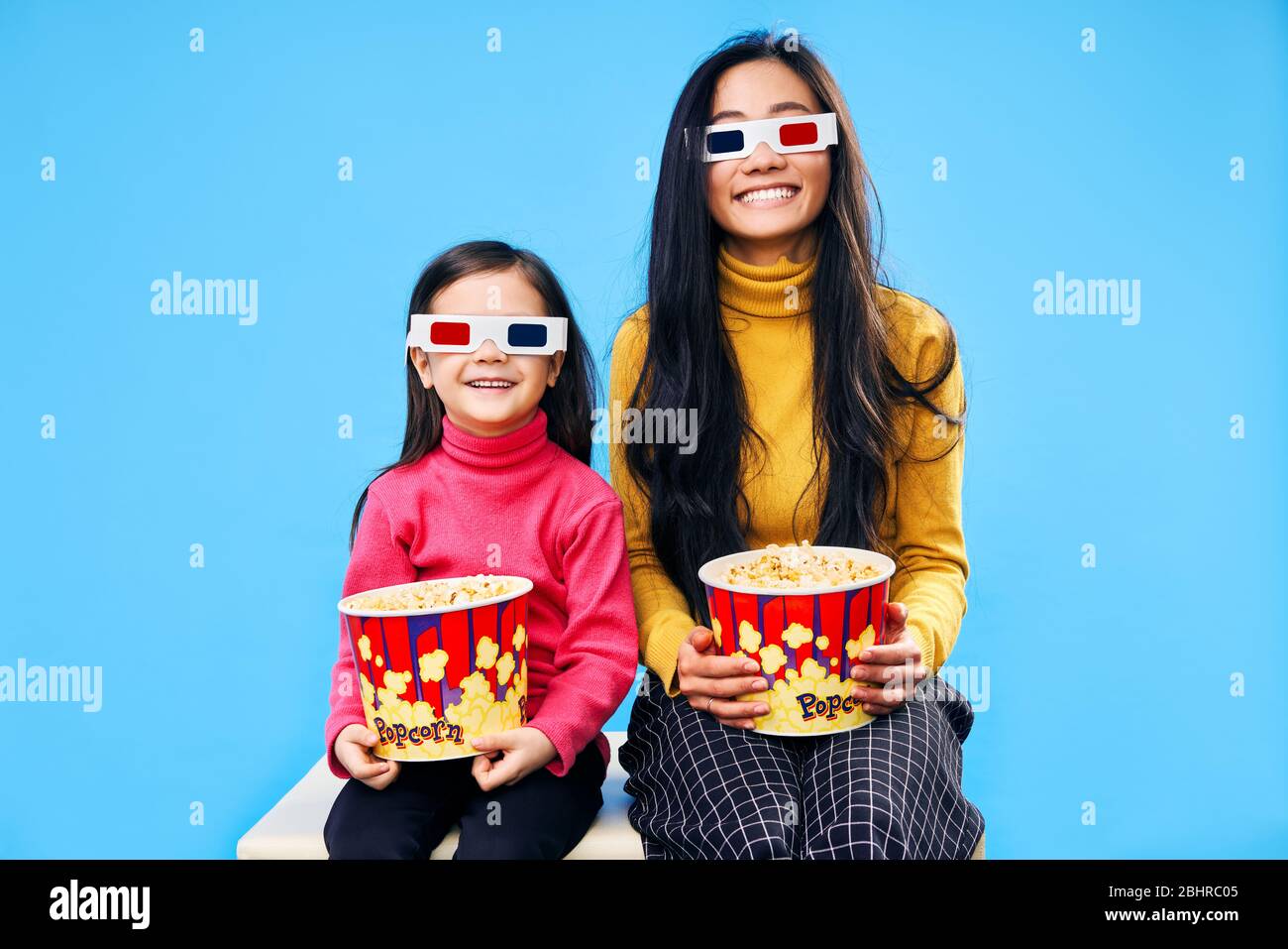 Happy smiling mother and her little daughter in 3D glasses with popcorn watching movie over blue background Stock Photo