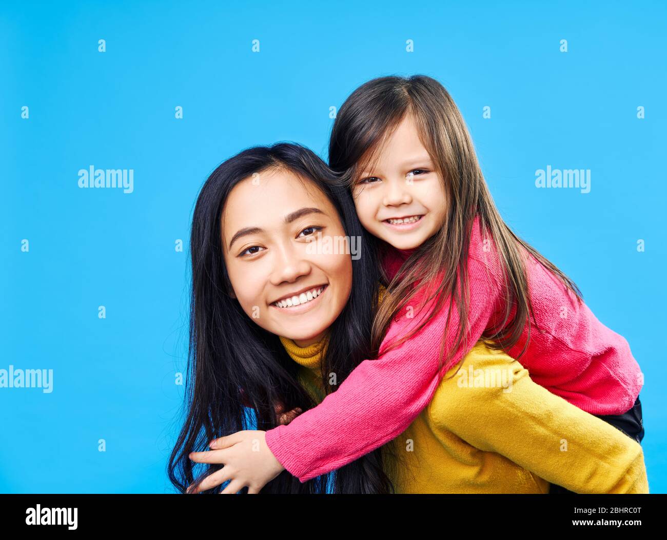 Happy mother and little daughter piggybacking together isolated on blue background. Family concept Stock Photo