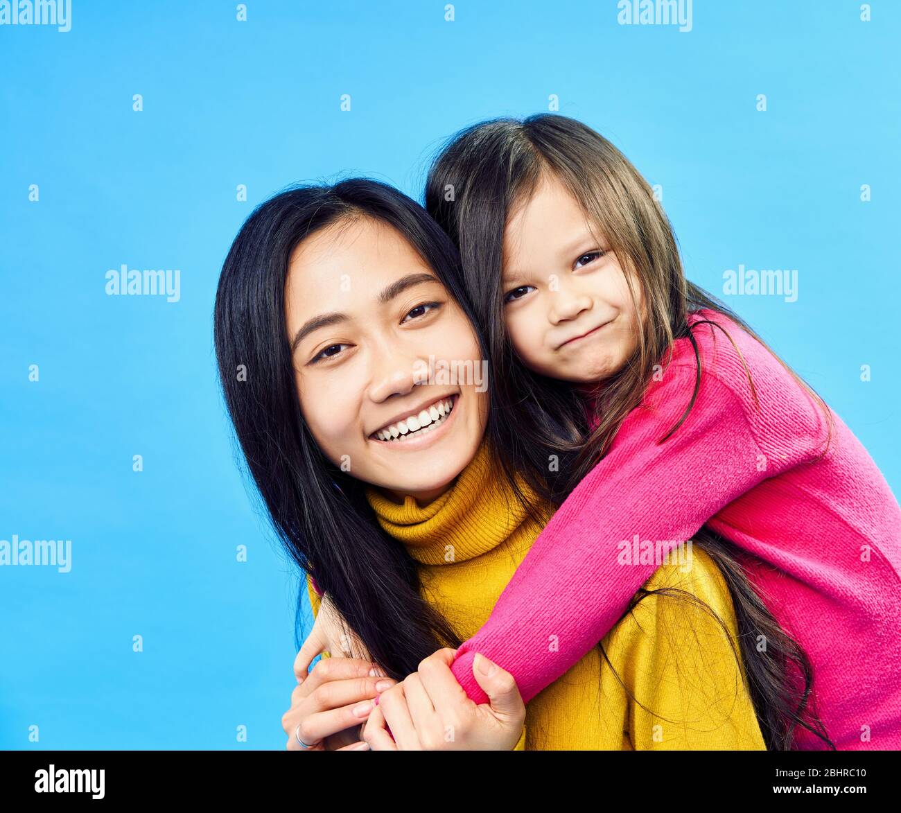 Happy mother and little daughter piggybacking together isolated on blue background. Family concept Stock Photo
