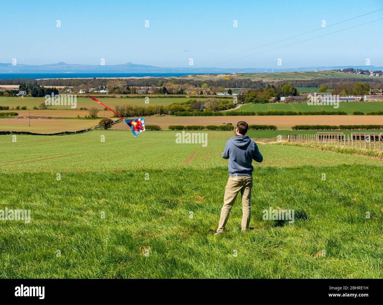 A man files a colourful kite on sunny day  with a view across the Firth of Forth to the twin peaks of Lomond Hills in Fife, East Lothian, Scotland, UK Stock Photo