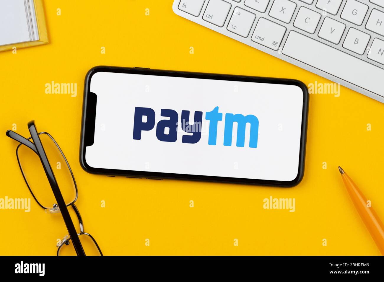A smartphone showing the PayTM logo rests on a yellow background along with a keyboard, glasses, pen and book (Editorial use only). Stock Photo
