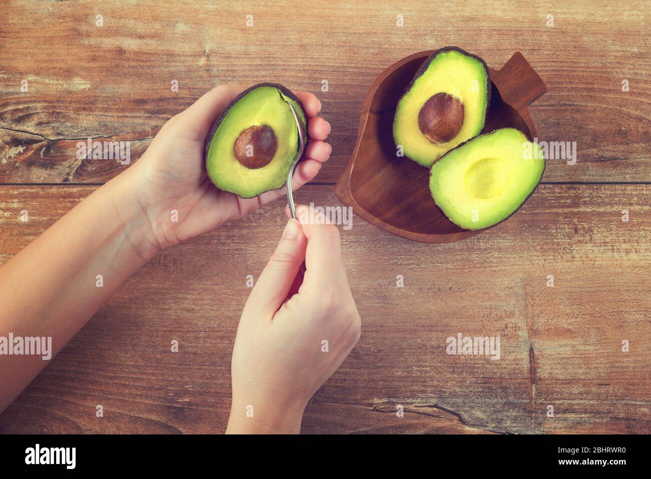 Female hands holding spoon and half of fresh ripe avocado. Woman peeling avocado. Green avocado on wooden table. Healthy eco food. Product of organic Stock Photo