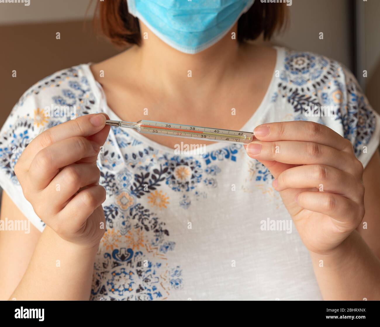 Coronavirus COVID-19 concept with female wearing mask holding thermometer with high temperature. Woman in domestic clothes, holds analogue medical Stock Photo