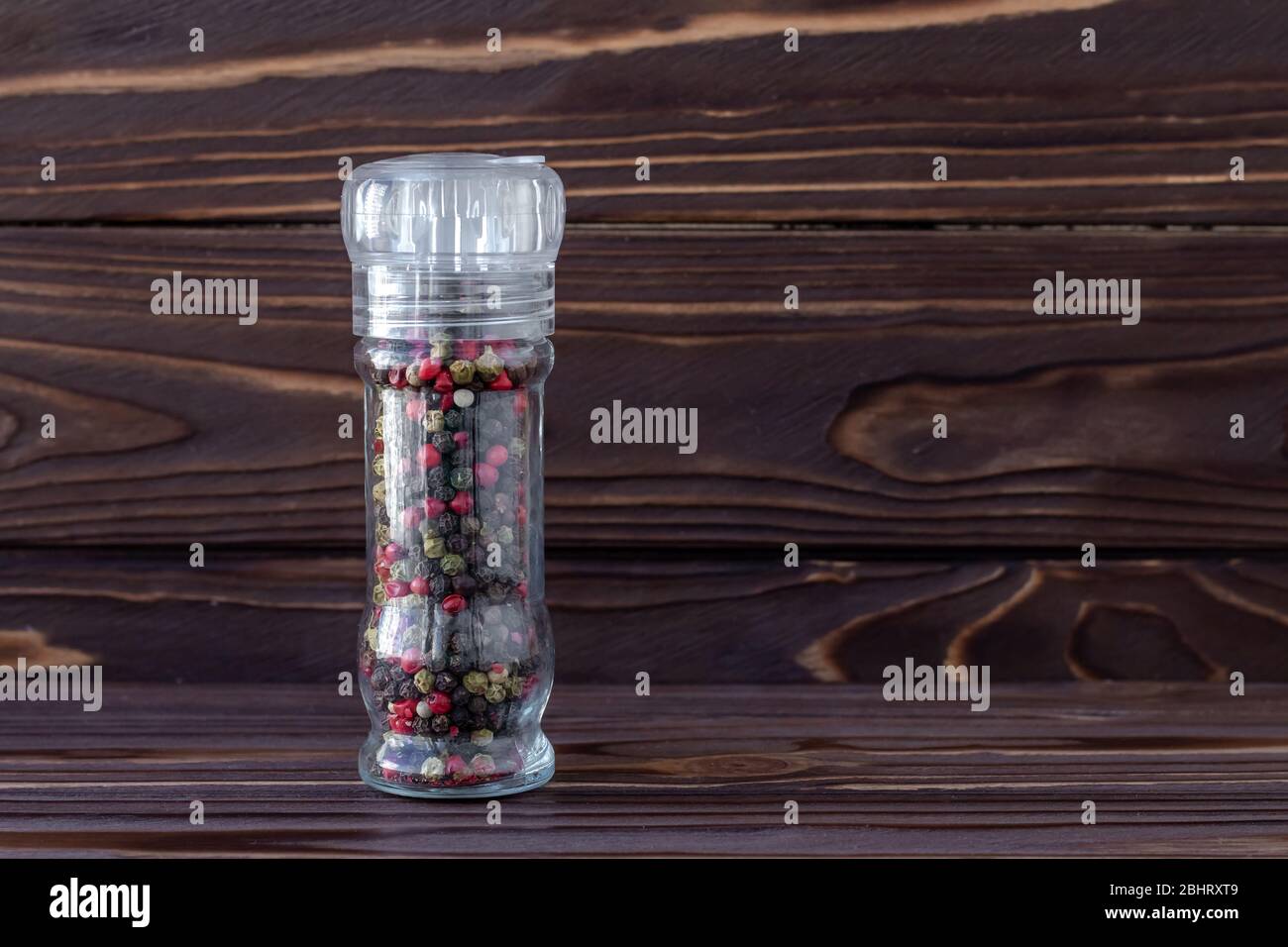 Colorful pepper peas in glass jar on dark brown wooden boards background, multicolored seeds of spicy peppercorn, hot red and black pepper in bottle. Stock Photo